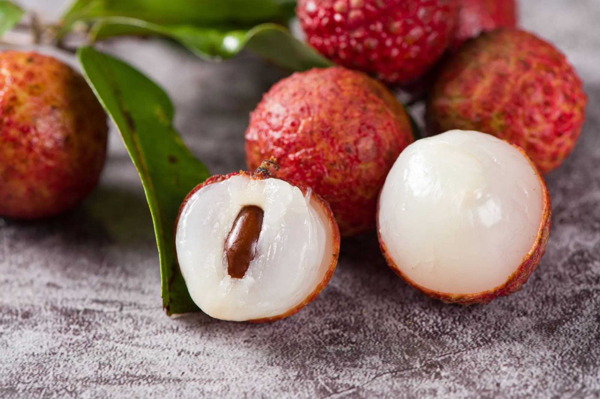 A lychee cut open to show the white flesh and dark seed, beside whole unpeeled fruit and a leaf