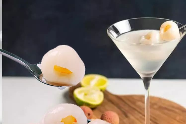 Peeled lychees in a glazed bowl, one lifted on a spoon, with a lychee cocktail alongside
