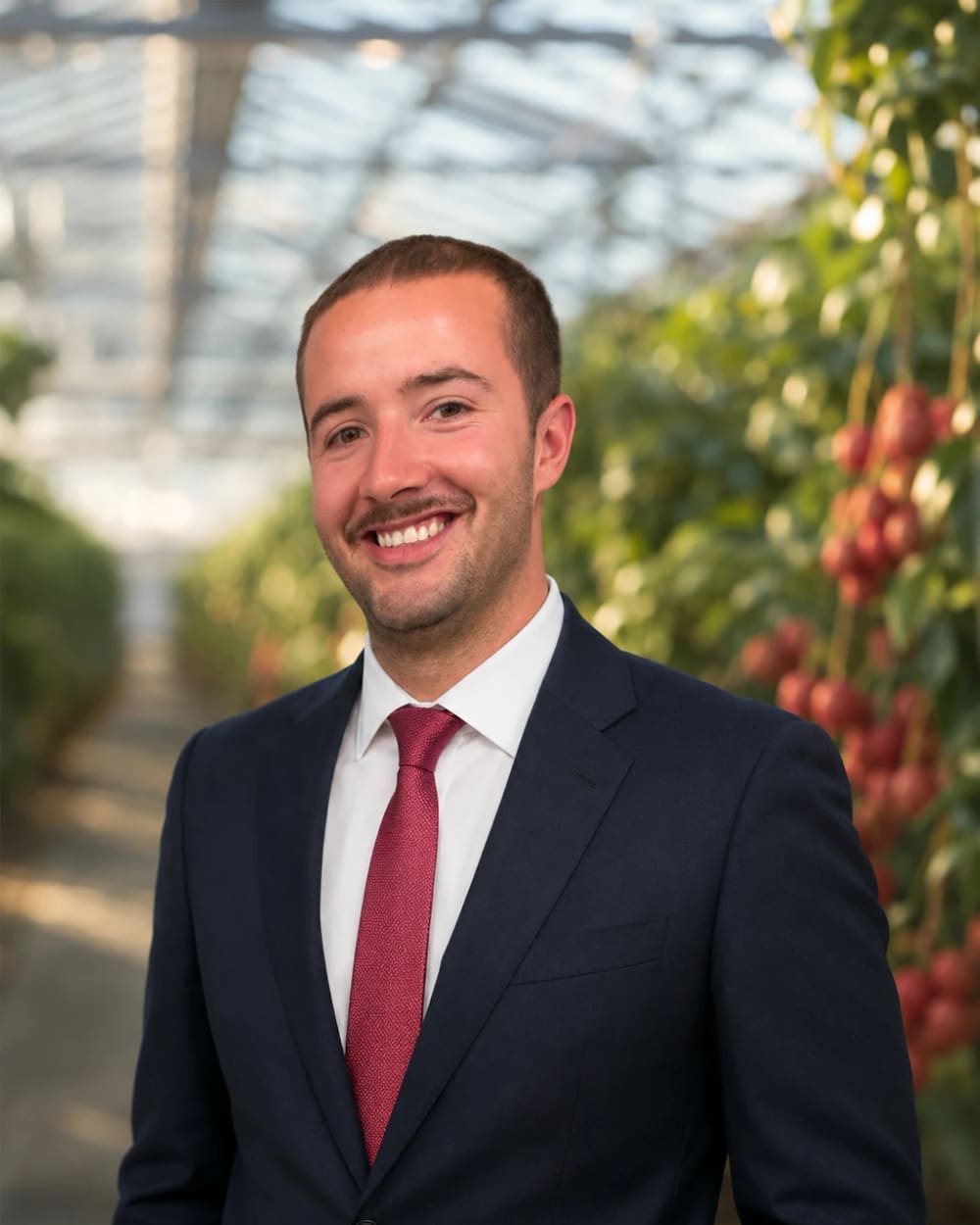 Julian in a navy suit and dark red tie, smiling, in one of the glasshouse rows with fruit on the trees behind him